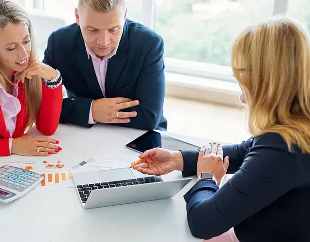 Un asesor financiero muestra datos en una computadora portátil a una pareja en una mesa blanca; se ven gráficos y una calculadora.