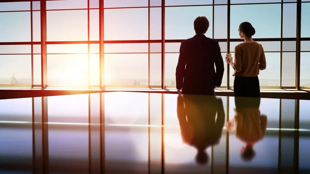Dos hombres de negocios se encuentran de pie mirando hacia afuera a través de una gran ventana, recortados contra una brillante puesta de sol que se refleja en el piso pulido.