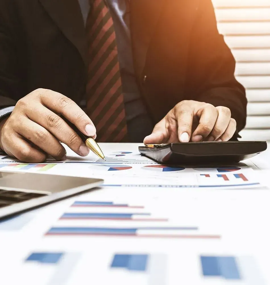 Persona con traje haciendo cálculos con una calculadora, apuntando con un bolígrafo a gráficos y datos financieros con una computadora portátil visible en el lado izquierdo.