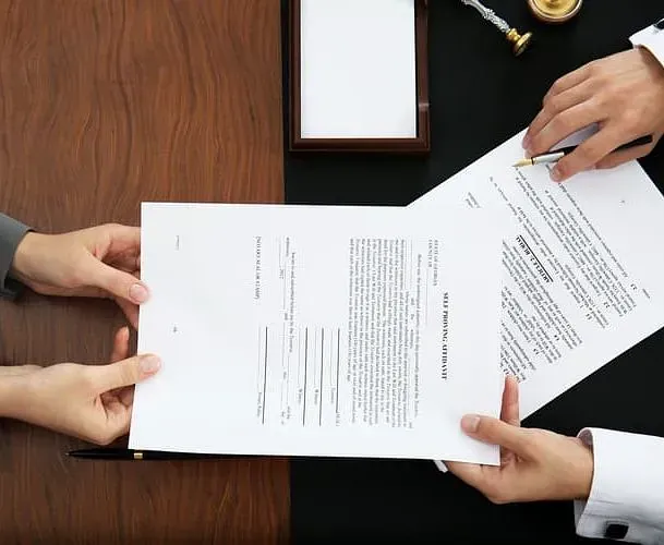 Dos personas intercambiando documentos a través de una mesa de madera, posiblemente durante una reunión legal o de negocios.