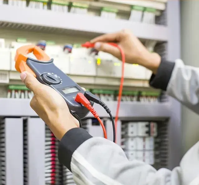 Electricista usando un multímetro para probar el cableado eléctrico de un panel de control. Lleva uniforme gris claro y sostiene el multímetro en las manos.