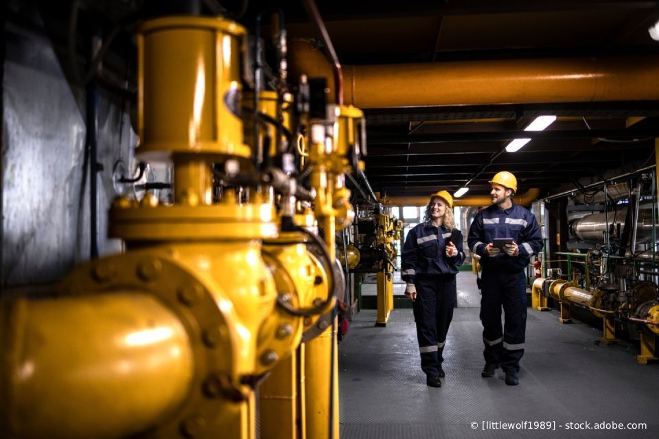 Zwei Arbeiter in Schutzkleidung gehen durch eine Industrieanlage mit gelben Rohren.