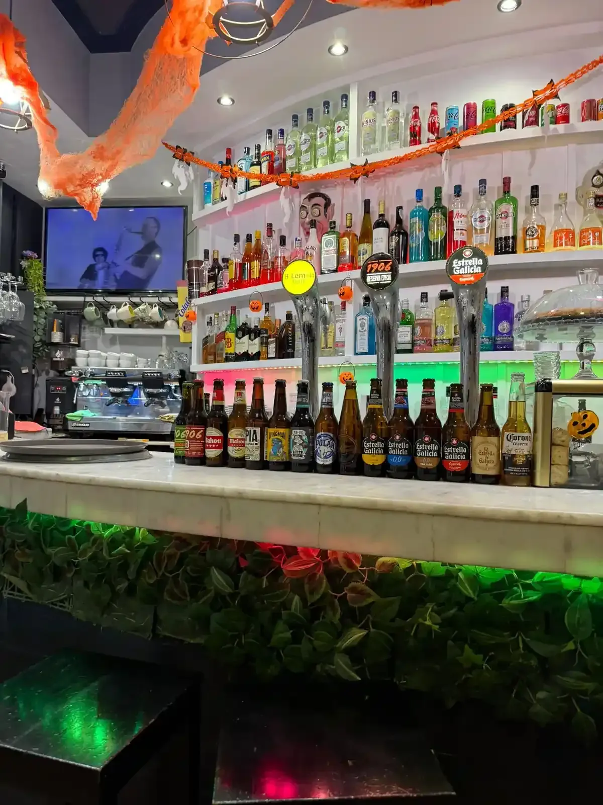 Interior de bar con botellas, grifos y molduras verdes brillantes. Televisión encendida.