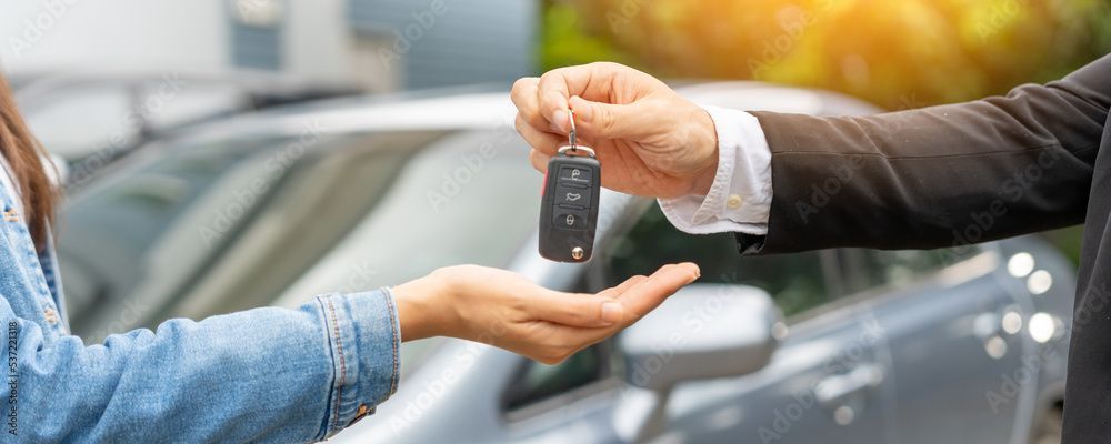 Übergabe von Autoschlüsseln neben einem geparkten Fahrzeug, im Hintergrund ist eine Zahlungskarte zu sehen.