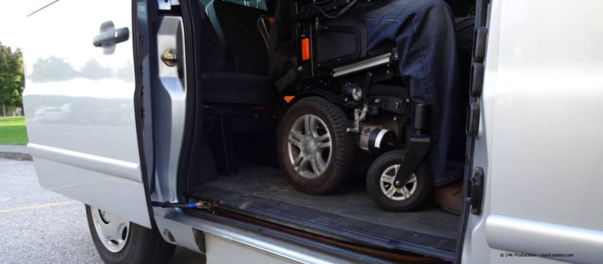 Eine Person im Rollstuhl sitzt in einem silbernen, barrierefreien Transporter mit geöffneter Schiebetür.