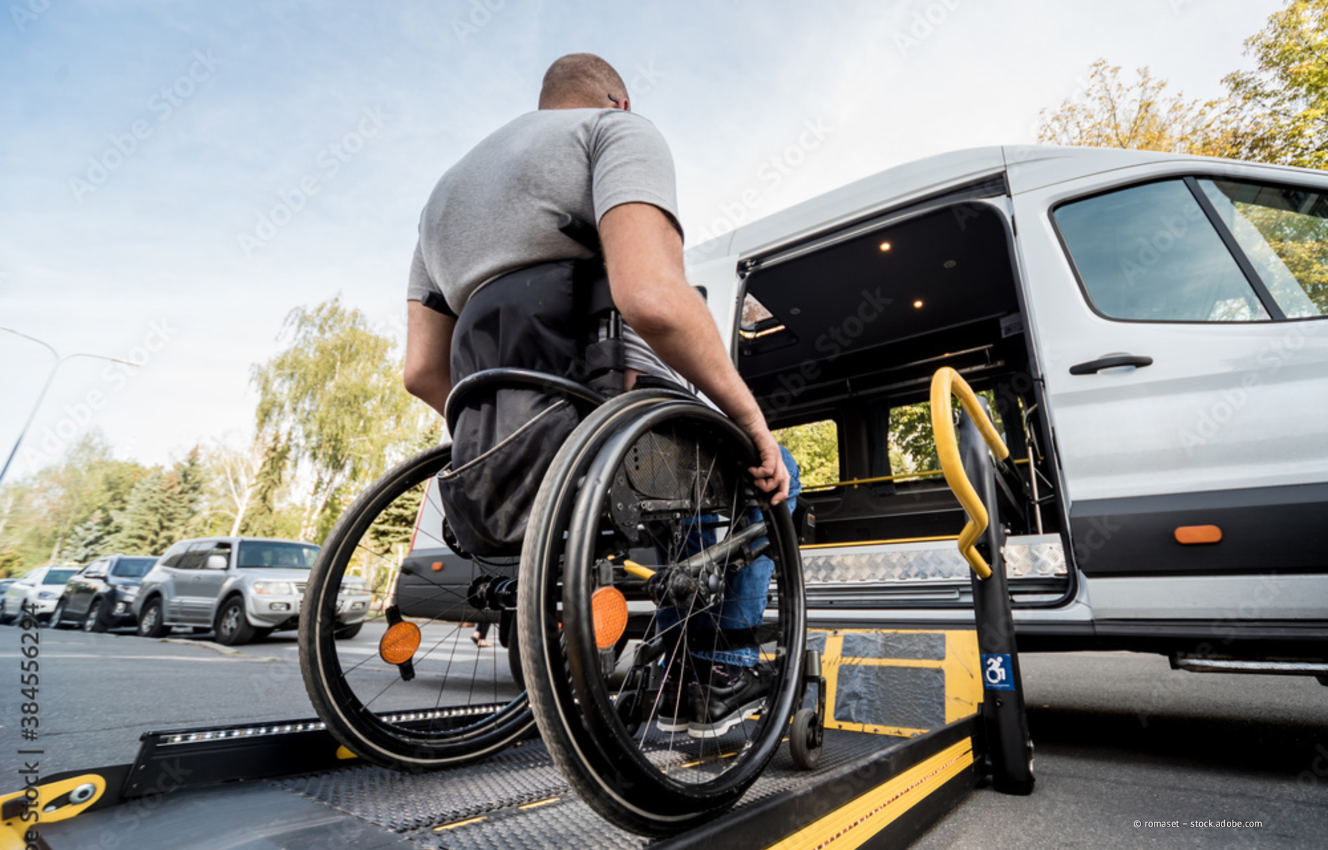Eine Person im Rollstuhl benutzt einen Lift, um in einen weißen, barrierefreien Kleinbus einzusteigen, der auf einer Straße in der Stadt geparkt ist.
