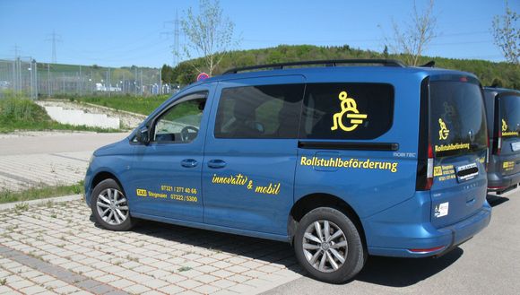 Ein silberner, rollstuhlgerechter Transporter, dessen automatische Rampe zu einer gepflasterten Fläche heruntergefahren ist.