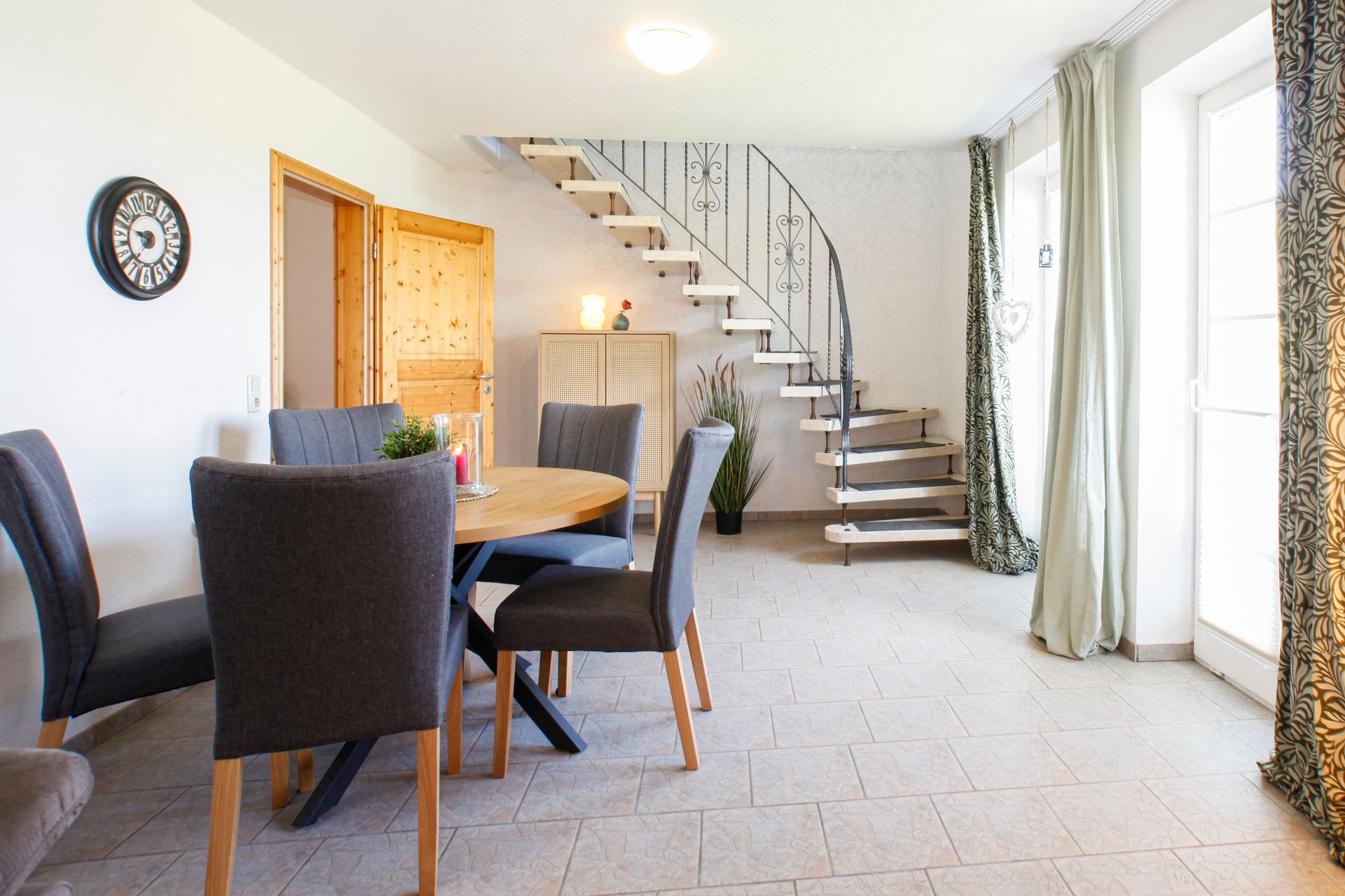 Helles Esszimmer mit dunklen Stühlen, Holztisch, geschwungener Treppe und großen Fenstern mit Vorhängen.
