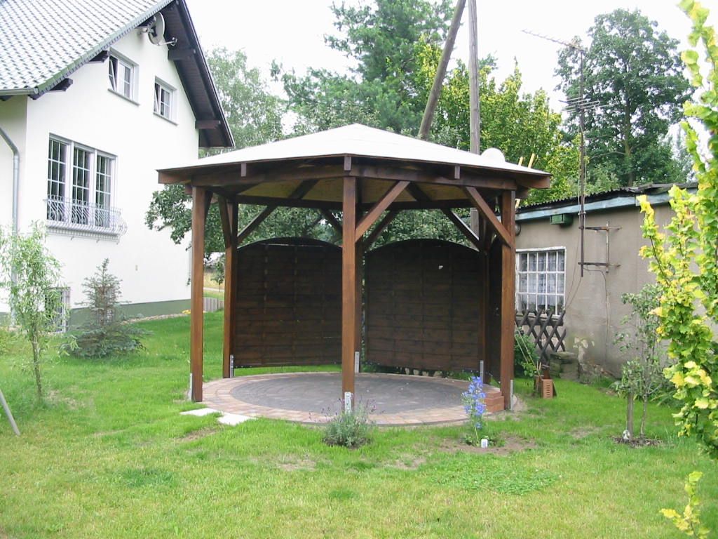 Ein hölzerner Pavillon in einem grasbewachsenen Hof, teilweise von einem braunen Zaun umgeben.