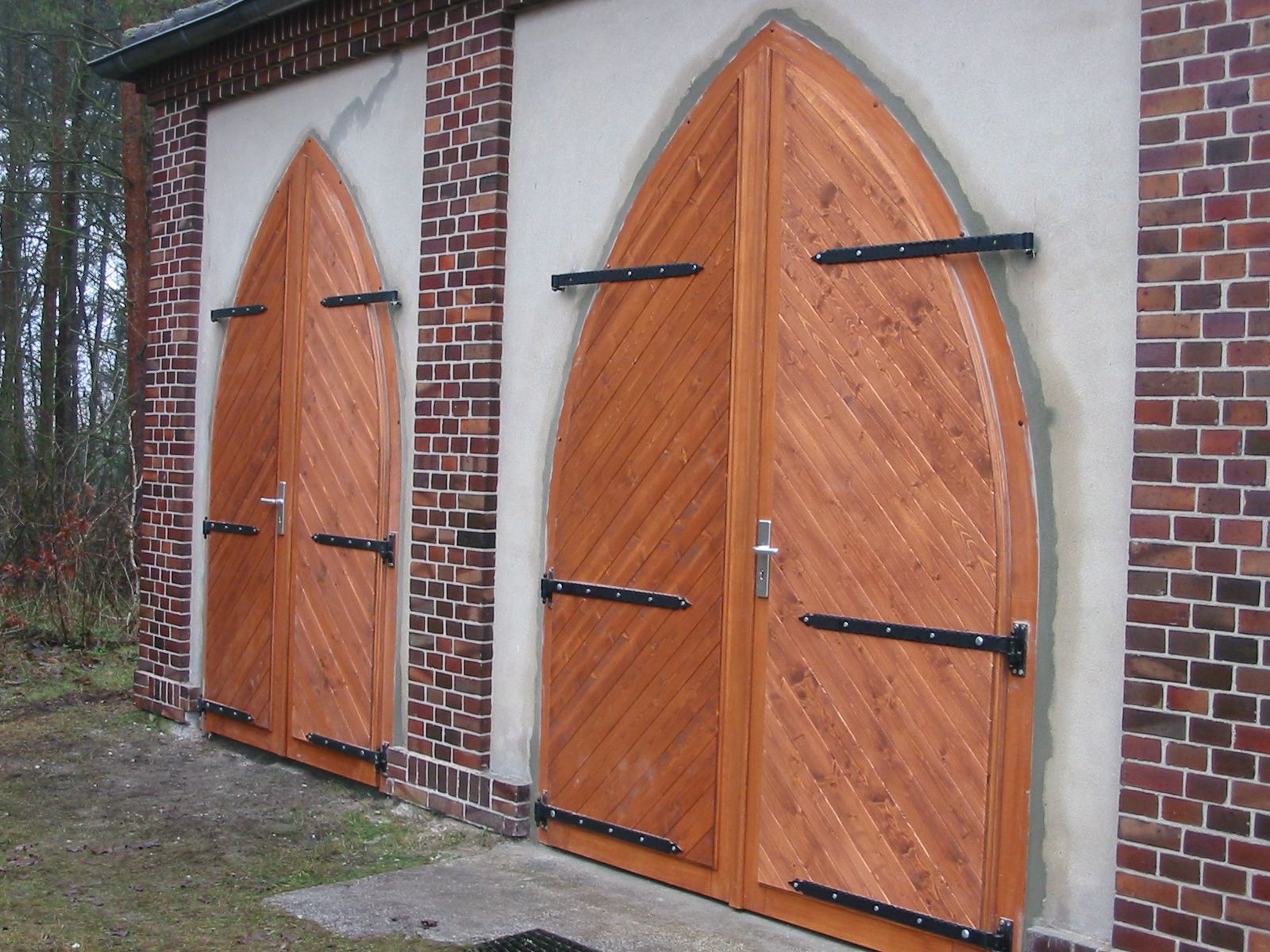 Zwei hölzerne Bogentüren mit schwarzen Beschlägen, eingelassen in eine Mauer aus Ziegeln und Stuck.