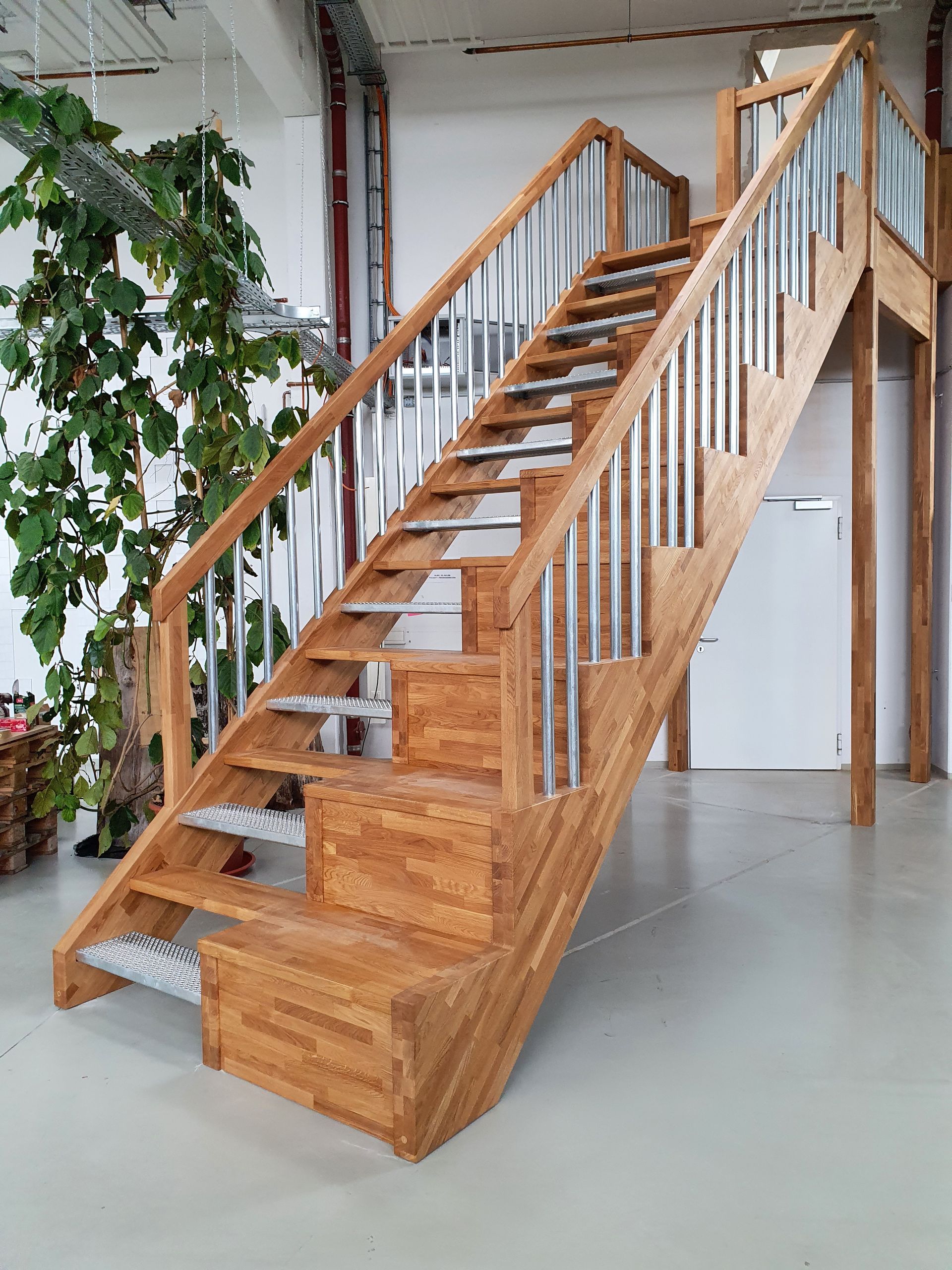 Holztreppe mit Metallgeländer in industriellem Ambiente, neben Grünpflanzen und einer weißen Tür.