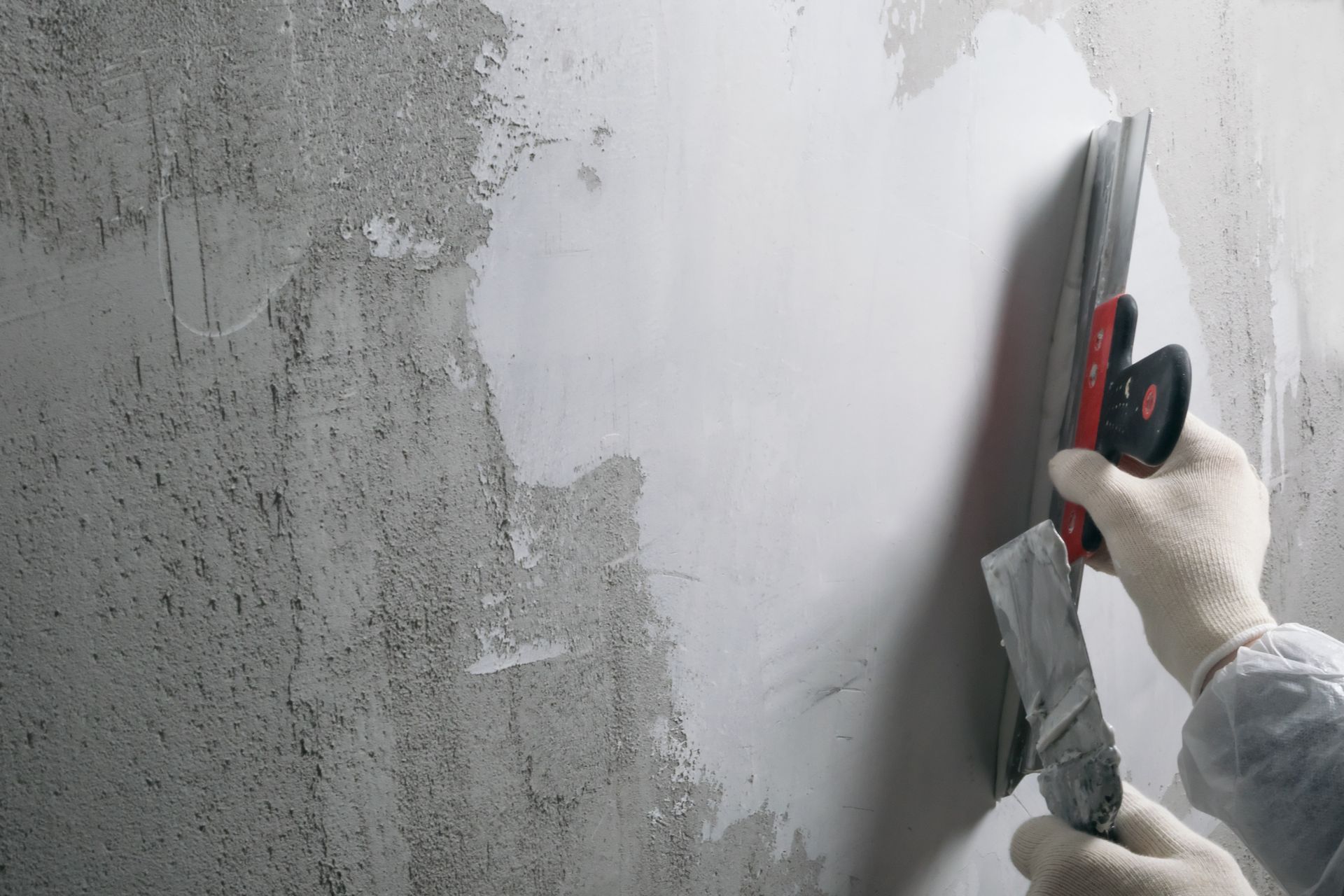 Eine Person trägt mit einer Kelle Gips auf eine Betonwand auf; weisser Gips, graue Wand, behandschuhte Hände.