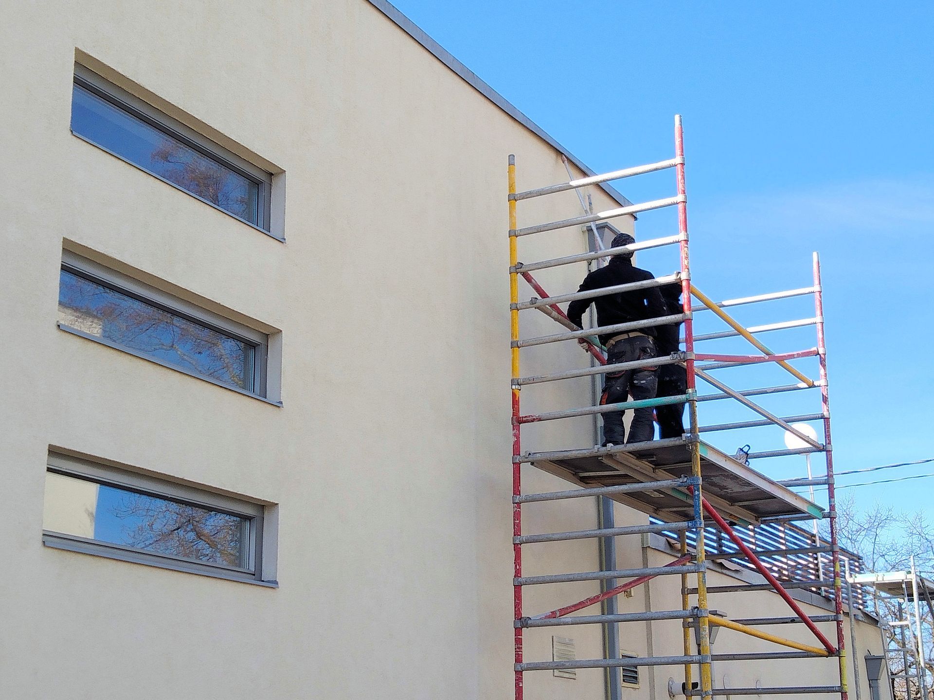 Un ouvrier sur un échafaudage peint la façade d’un bâtiment, à côté de fenêtres étroites.
