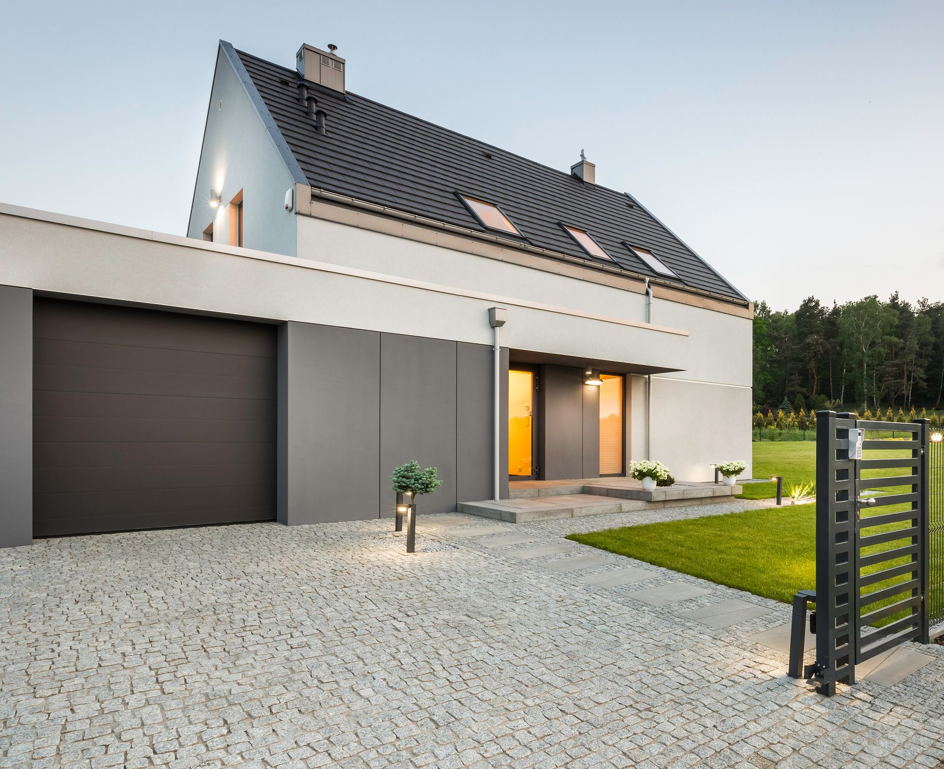 Extérieur d’une maison moderne au crépuscule, avec un toit sombre, une façade blanche, un garage, une allée en gravier et une pelouse