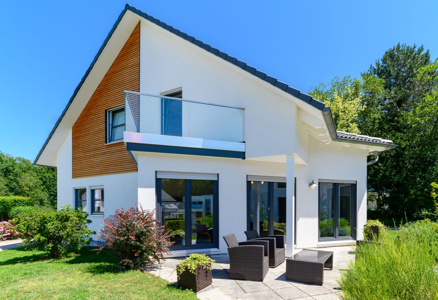 Maison blanche moderne avec balcon, grandes fenêtres et chaises de jardin dans un jardin verdoyant et ensoleillé