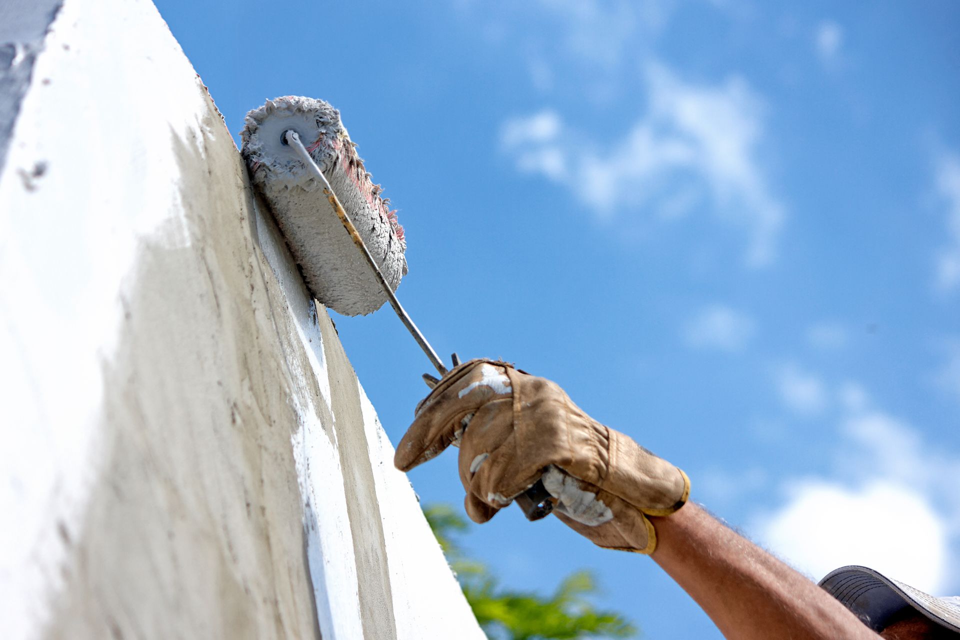 Une main gantée peint un mur blanc au rouleau, sur fond de ciel bleu