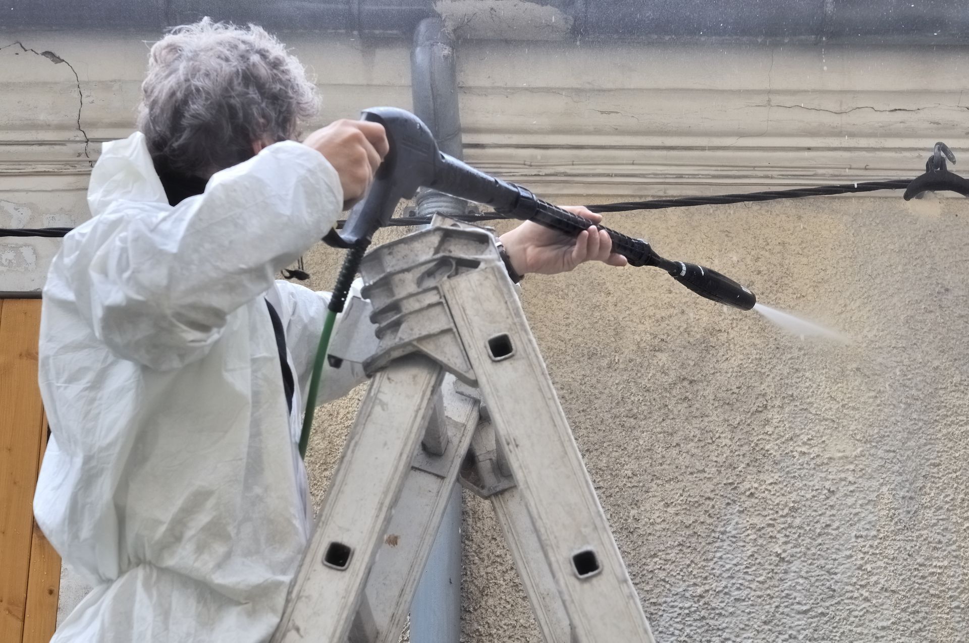 Personne nettoyant un mur au nettoyeur haute pression depuis une échelle, avec un tuyau et une lance de pulvérisation