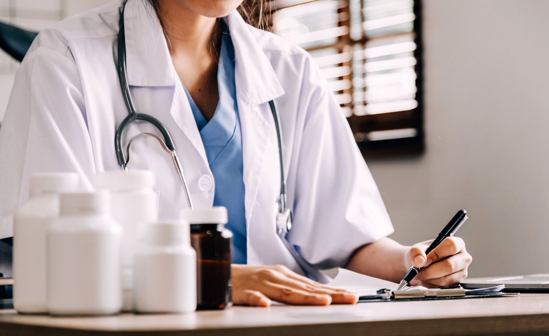 Una doctora está sentada en un escritorio escribiendo en un portapapeles.