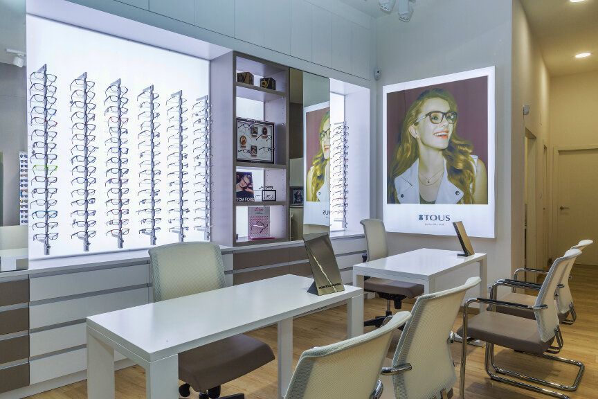 Interior de una óptica con vitrinas blancas que contienen gafas, pequeños escritorios blancos y un póster de una mujer con gafas.
