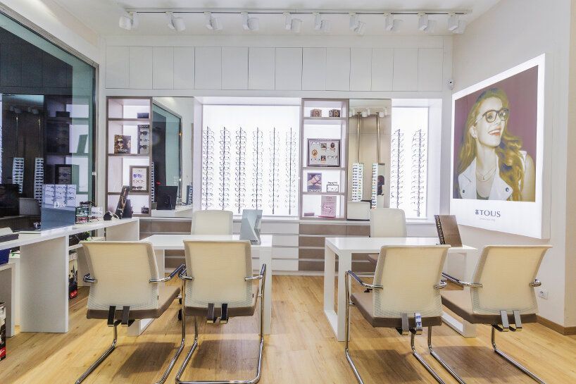 Interior de tienda de óptica con expositores de gafas, mesas blancas y sillas.