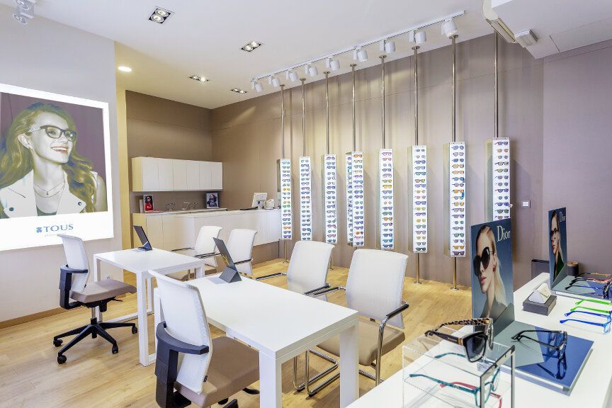 Interior de una tienda de gafas. Vitrinas con monturas, mesas blancas, sillas y carteles publicitarios.