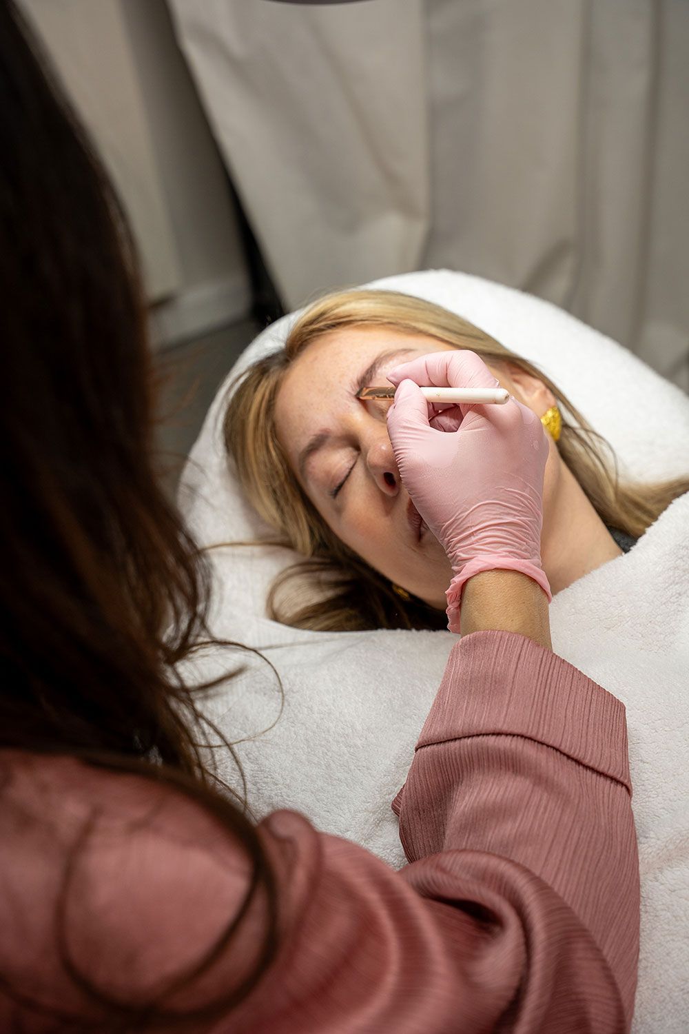Eine Frau lässt sich von einer Frau mit rosa Handschuhen die Augenbrauen machen.