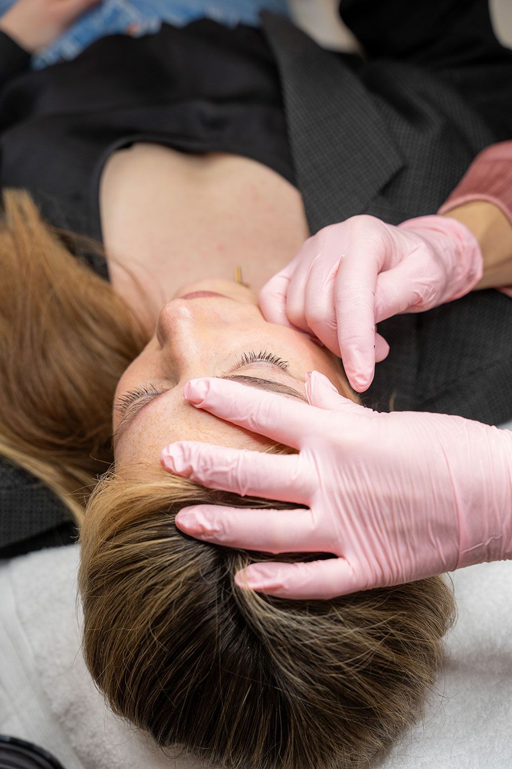 Eine Frau liegt mit geschlossenen Augen auf dem Bauch, während eine Person mit rosa Handschuhen ihr Gesicht berührt.