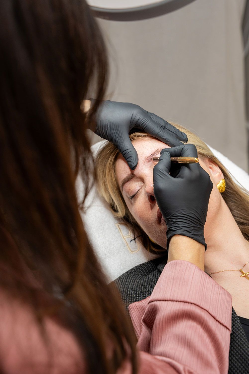 Eine Frau lässt sich von einer Frau mit schwarzen Handschuhen die Augenbrauen machen.