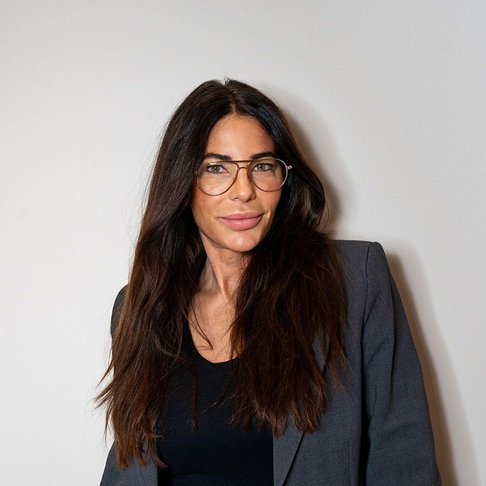 Eine Frau mit Brille und grauer Jacke steht vor einer weißen Wand.