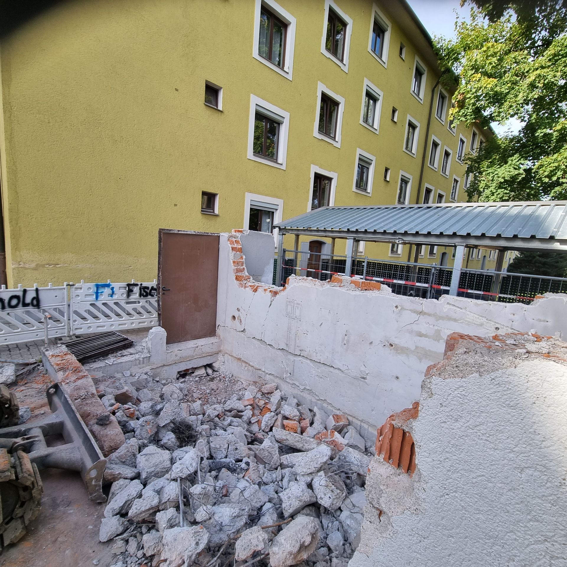 Abgerissene Ziegelmauer mit Schutt, braune Tür, gelbes Gebäude im Hintergrund.