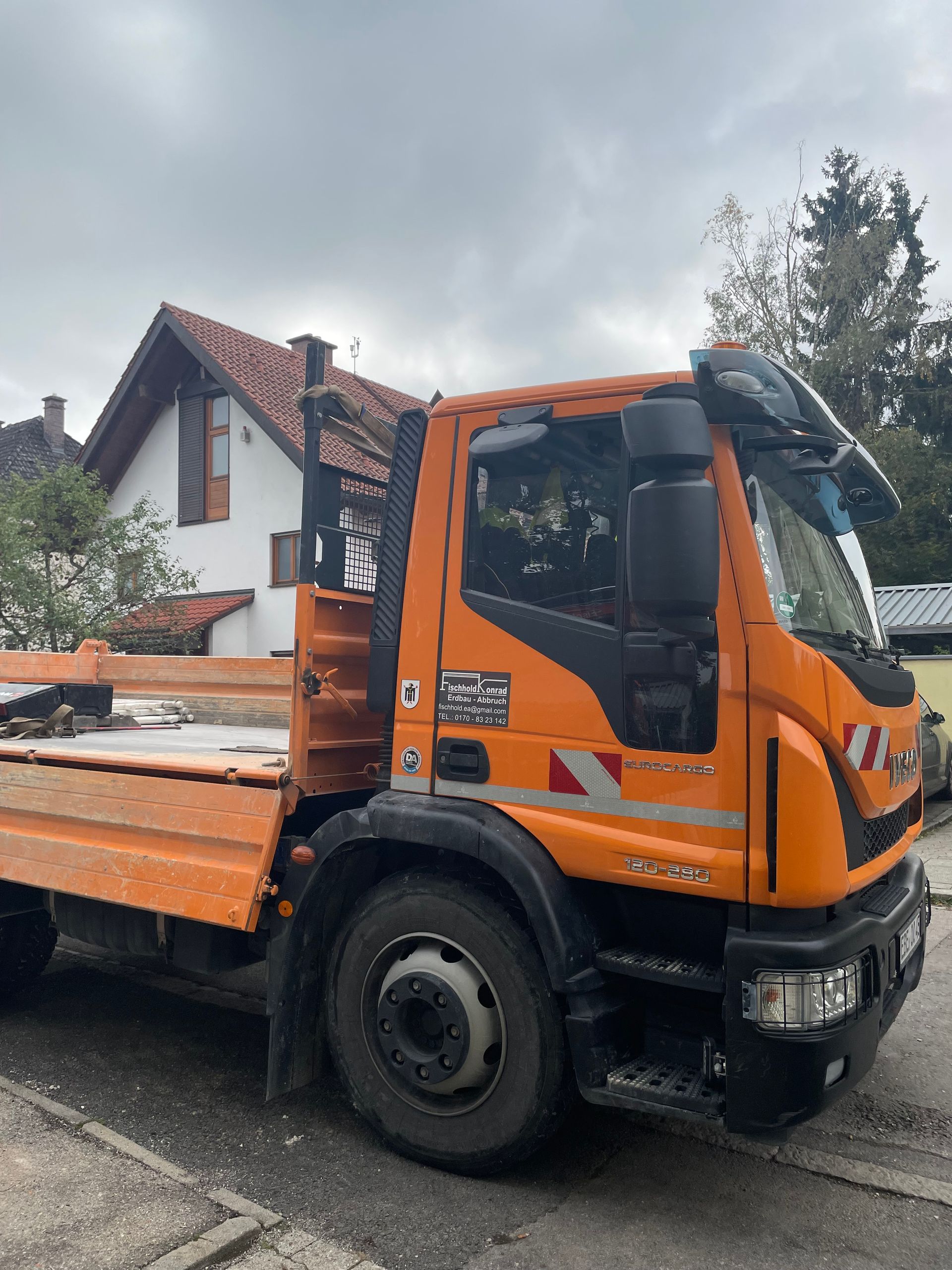 Orangefarbener Pritschenwagen, der vor einem Haus geparkt ist, im Hintergrund ist ein bewölkter Himmel zu sehen.