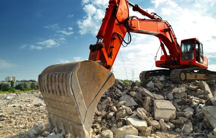 Ein gelber Bagger zerstört ein Gebäude, im Hintergrund ist blauer Himmel.