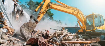 Ein gelber Bagger zerstört ein Gebäude und wirbelt unter blauem Himmel Staub und Schutt auf.