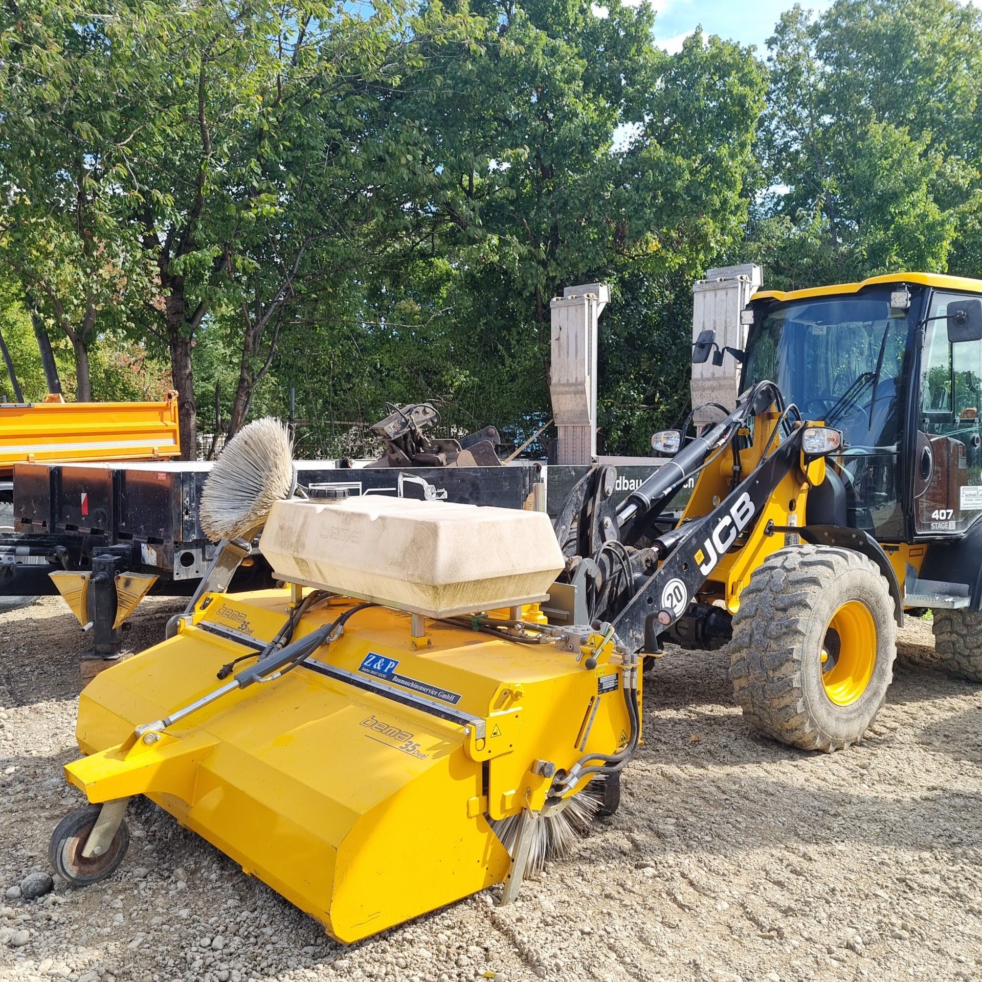 Gelber JCB-Kompaktradlader mit Kehrmaschinenaufsatz, im Freien.