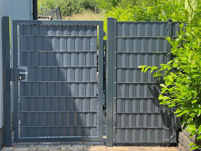 Graues Metalltor und Zaun mit vertikalen Paneelen, in einem Garten mit viel Grün.