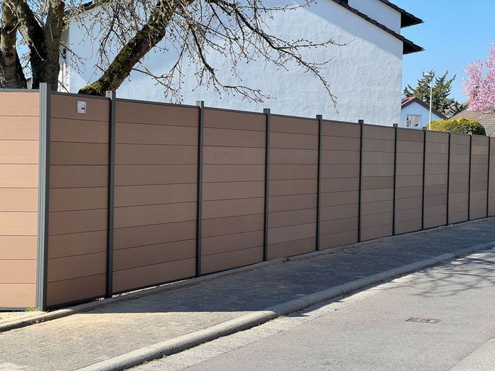 Brauner Verbundzaun grenzt an eine graue Schotterstraße, im Hintergrund ein weißes Haus.