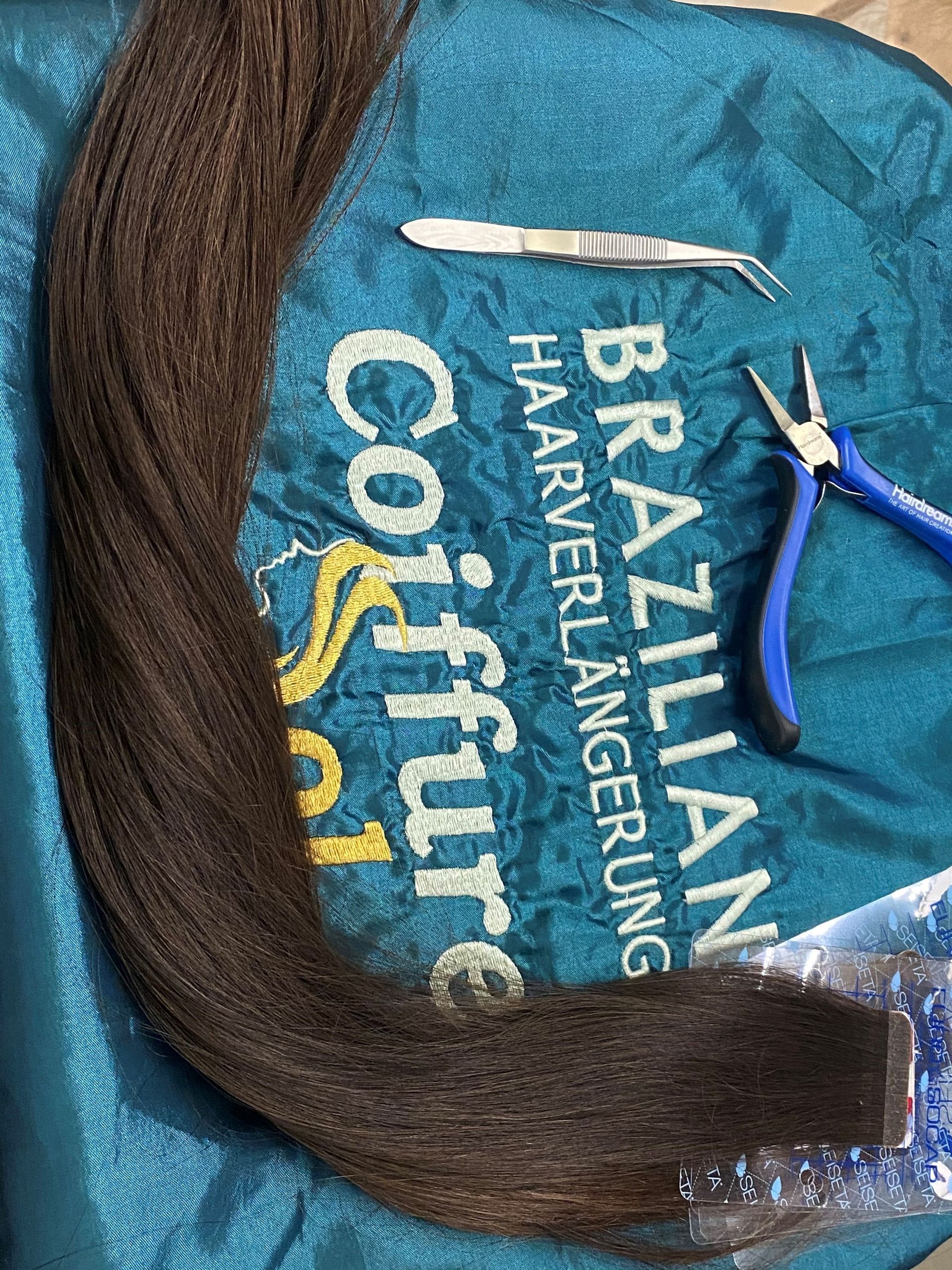 Braune Haarverlängerungen, Zange, Pinzette und ein blauer Beutel mit der Aufschrift „Brazilian Haarverlängerung“.