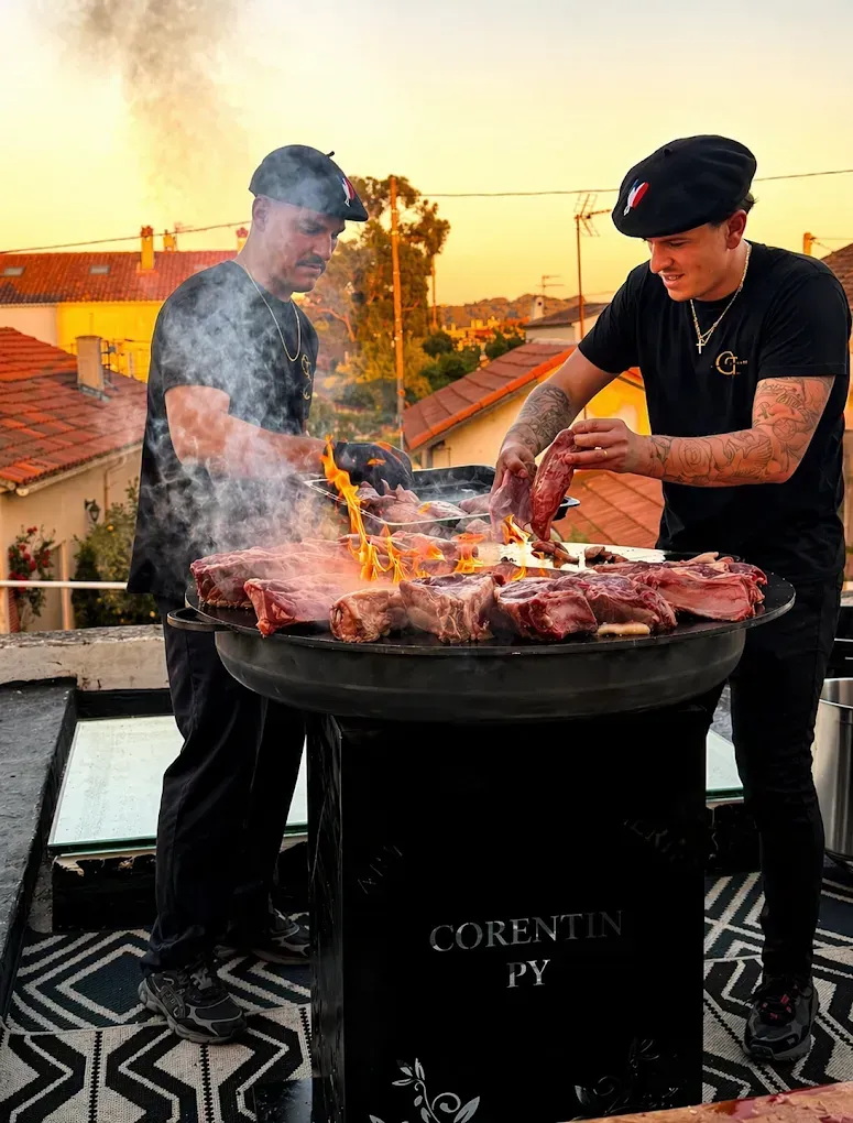 Deux chefs font griller de la viande sur un toit au coucher du soleil, avec de la fumée au-dessus d'un grand barbecue.