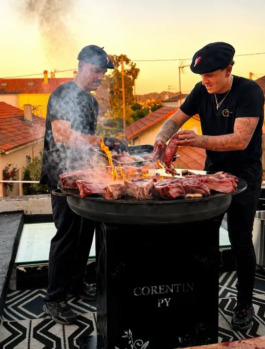 Deux personnes font griller de la viande sur un toit, au coucher du soleil, tandis que la fumée s'élève d'un grand brasero.