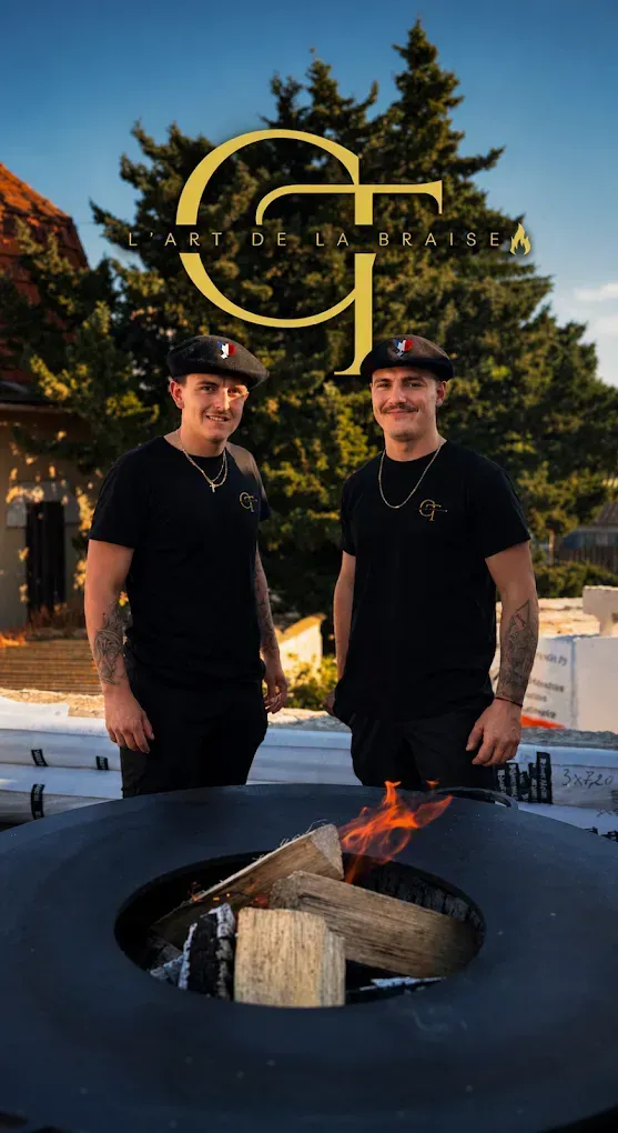 Deux chefs en uniforme noir se tiennent près d'un foyer circulaire où crépitent des flammes, dans un cadre extérieur.
