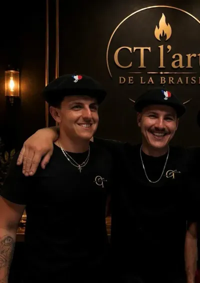 Deux personnes souriantes, vêtues de tee-shirts et de bérets noirs, posent bras dessus bras dessous devant une enseigne.