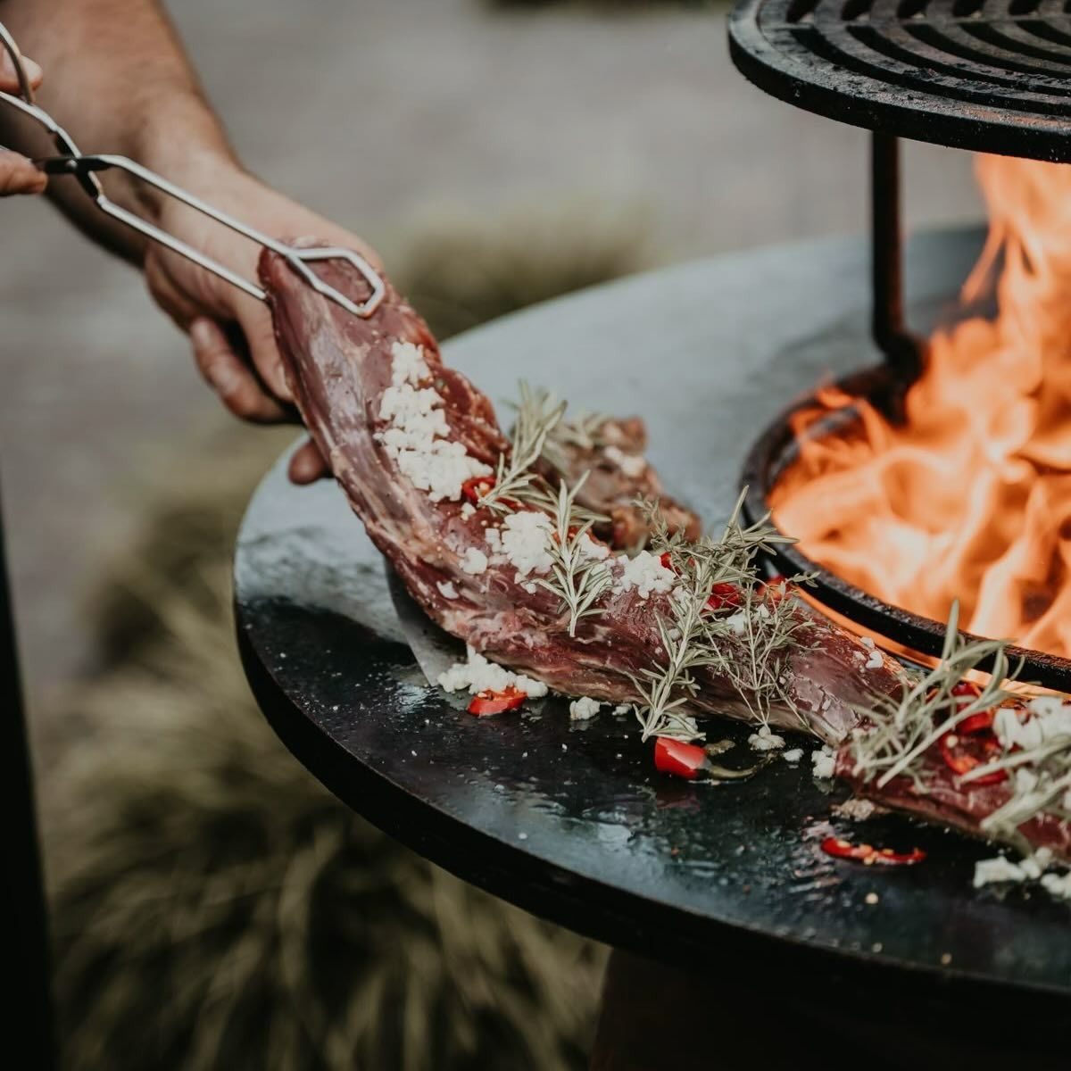 Déposer à la main d'une pièce de viande assaisonnée sur un gril de foyer extérieur avec des flammes visibles.