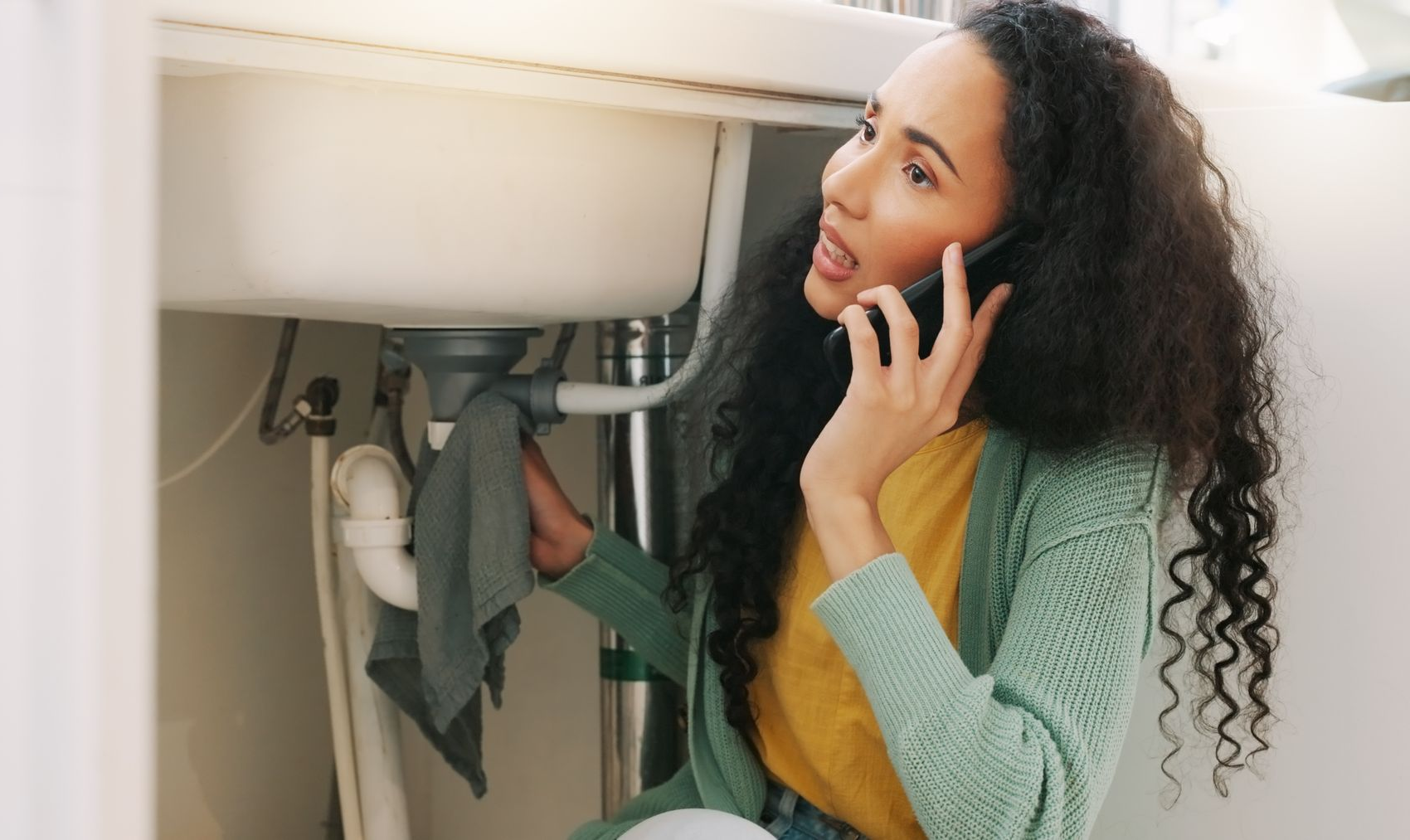 Une personne parle au téléphone tout en essuyant le dessous d'un évier de cuisine avec un chiffon.