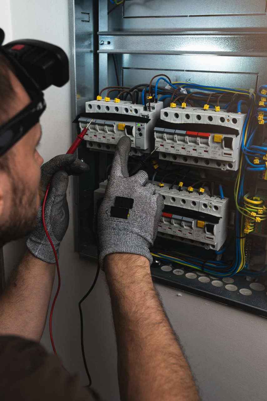 Un électricien teste les circuits d'un tableau électrique. Il porte des lunettes de sécurité et des gants.
