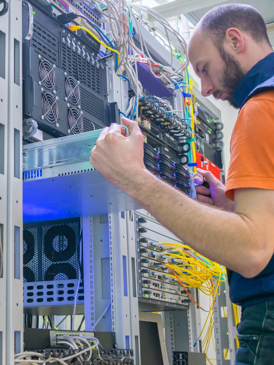 Un homme travaille sur une baie de serveurs, entouré de câbles. Il porte une chemise orange et un gilet bleu.