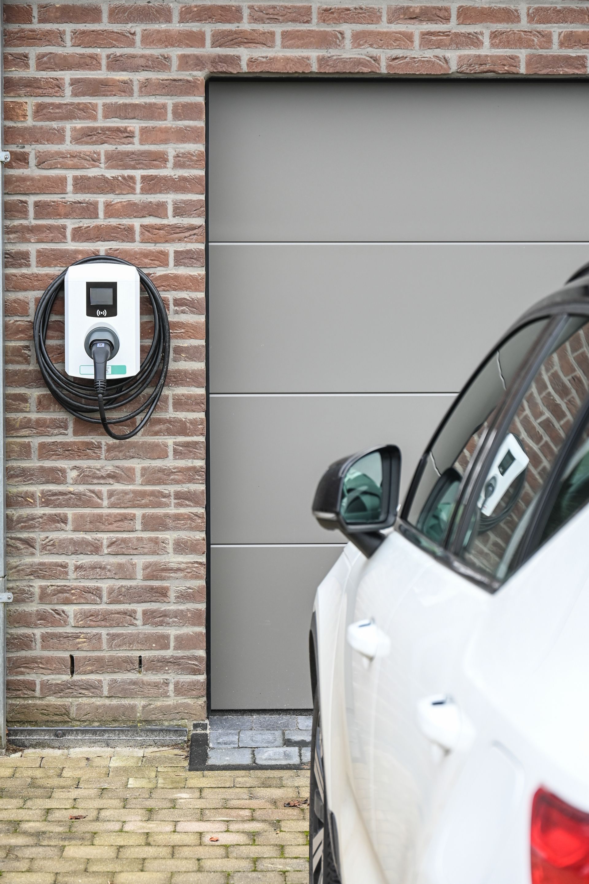 Voiture électrique en charge à côté d'un garage ; borne de recharge sur un mur de briques, voiture blanche visible.