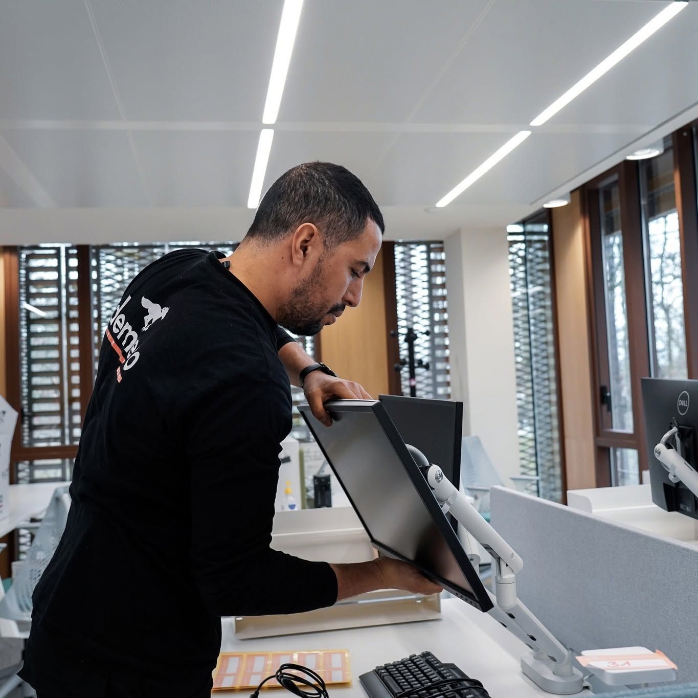 Un homme ajuste un écran d’ordinateur sur un bureau moderne, sous un éclairage vif au plafond