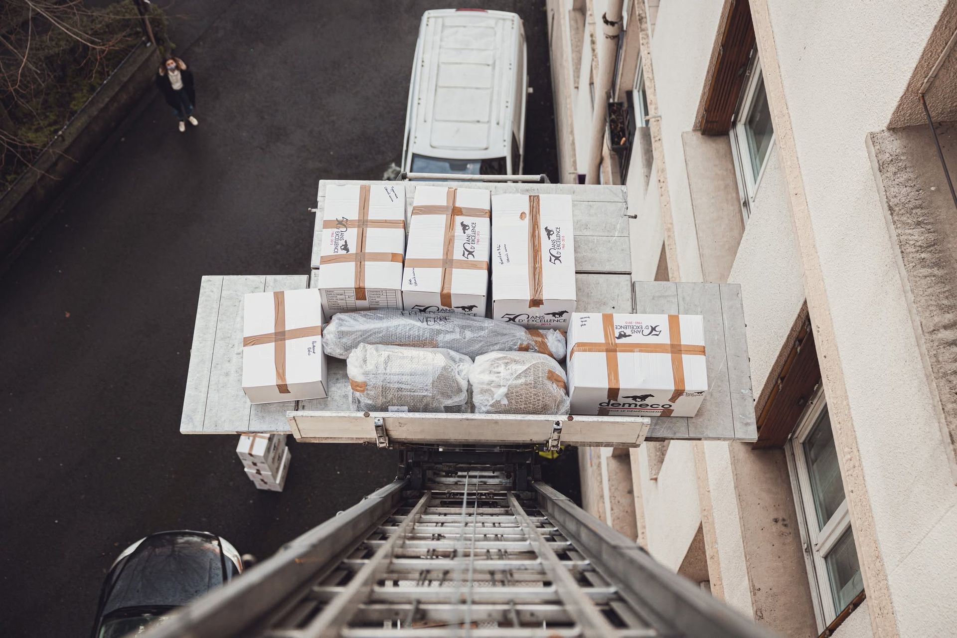 Un ouvrier sur une échelle hisse une palette de cartons sanglée, le long d’un mur de bâtiment