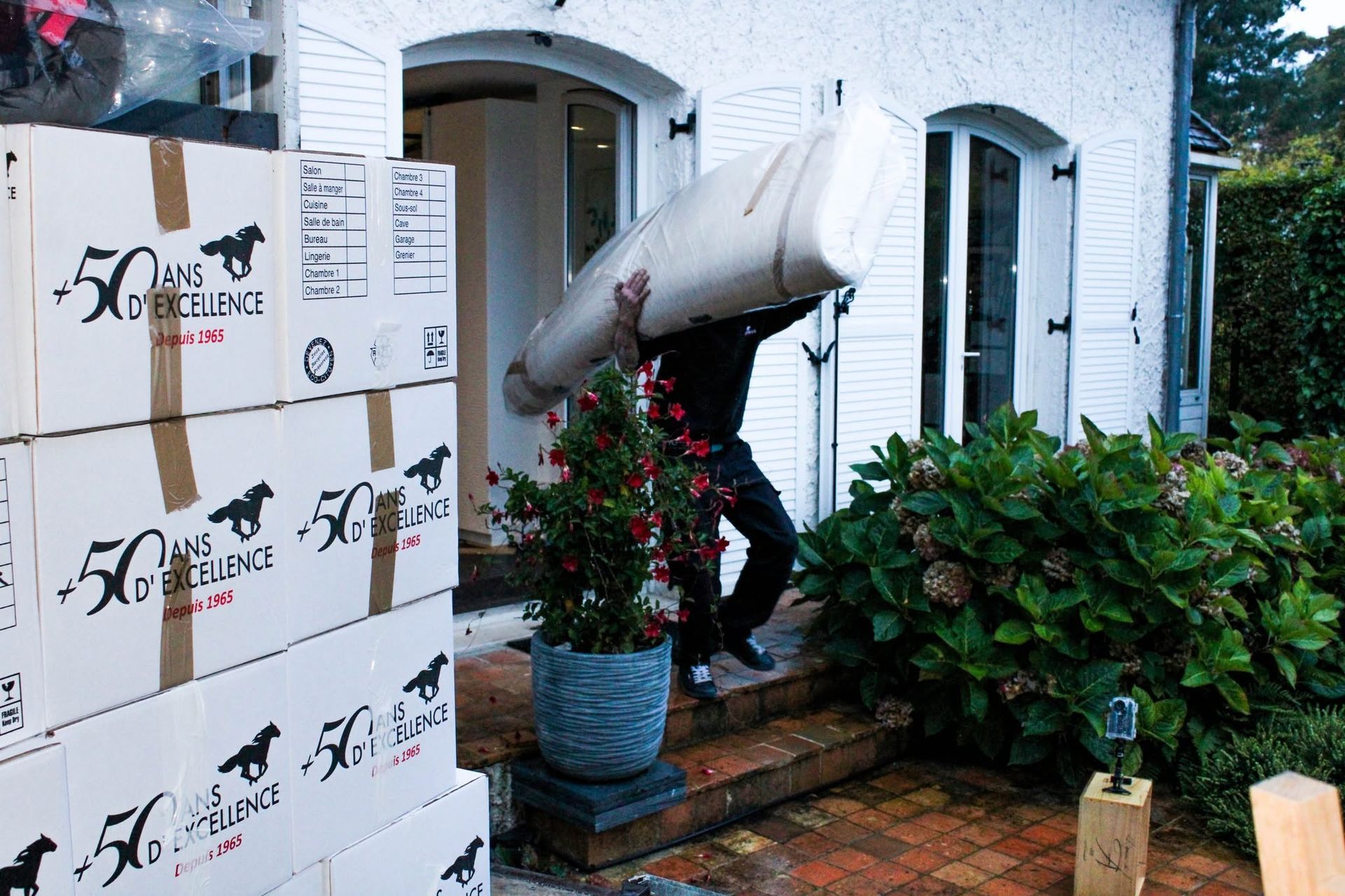 Des cartons empilés à côté de l’entrée d’une maison blanche, avec des fleurs en pot et une enseigne suspendue sur le porche