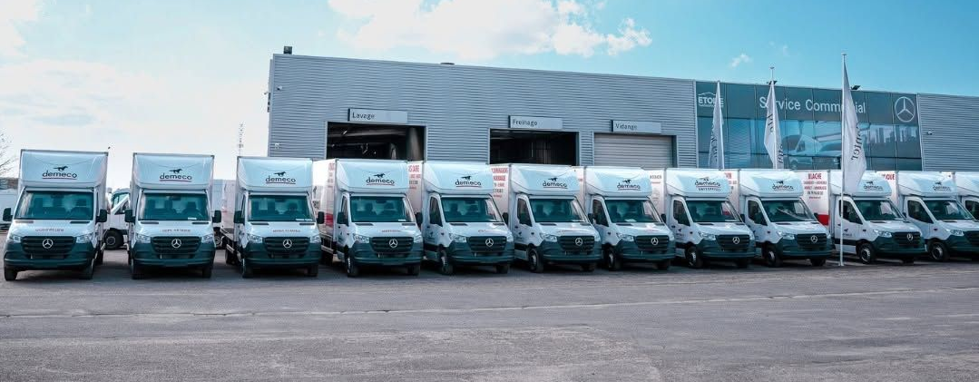 Rangée de camionnettes de livraison blanches garées devant un grand entrepôt sous un ciel nuageux